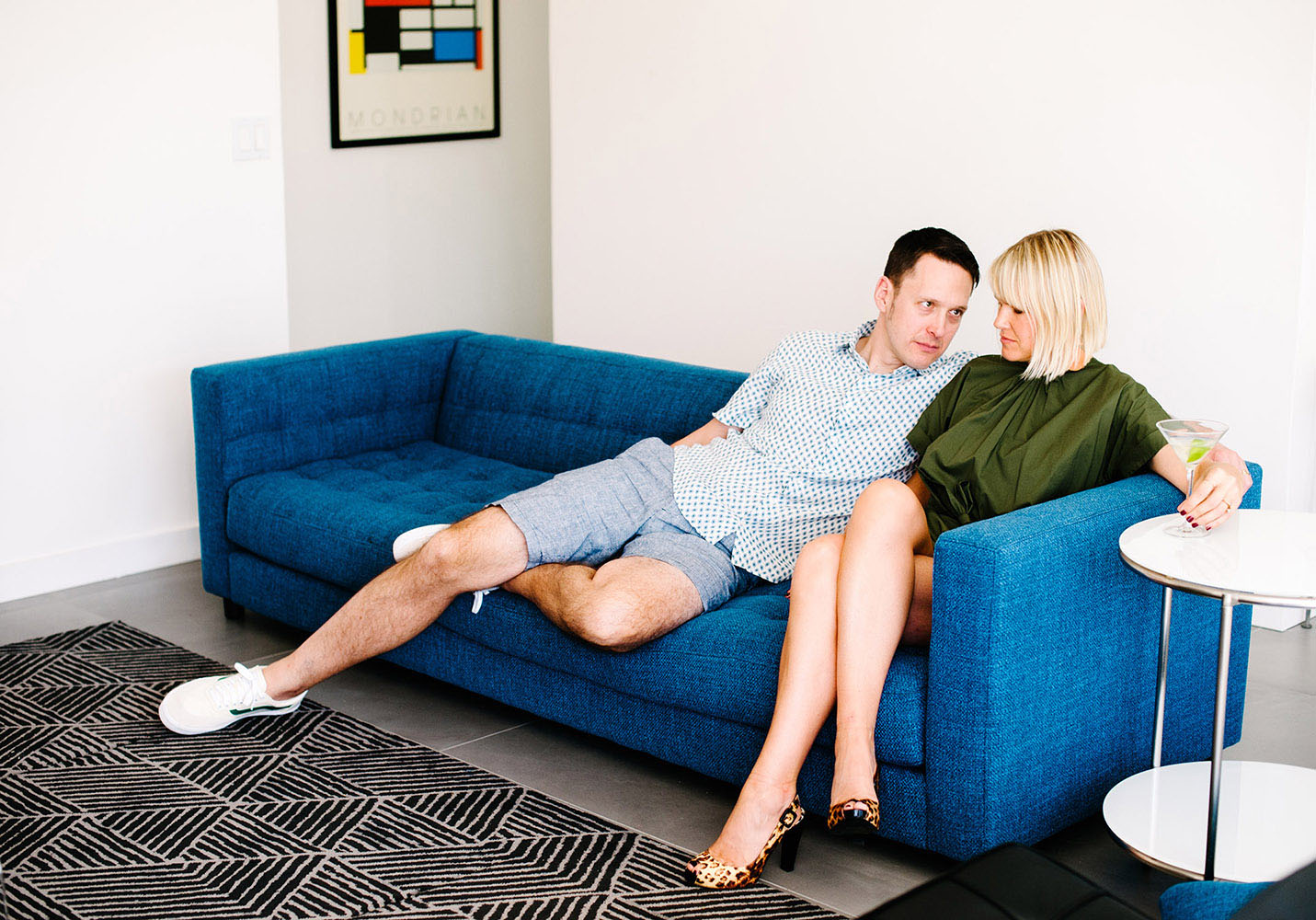 woman in green jumper with leopard print heels with martini on blue couch with guy in white shirt with blue dots and blue shorts and Mondrian print mid century modern furniture