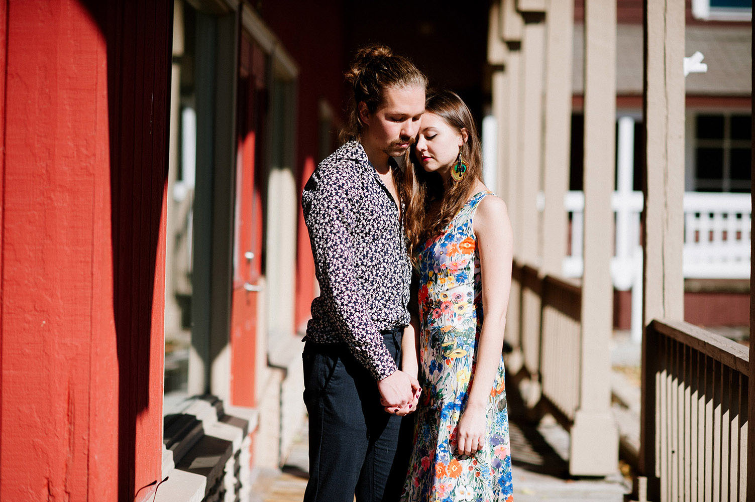 white male with facial hair and man bun with blue print shirt and female with long brown hair, peacock feahter earrings and floral print dress embracing