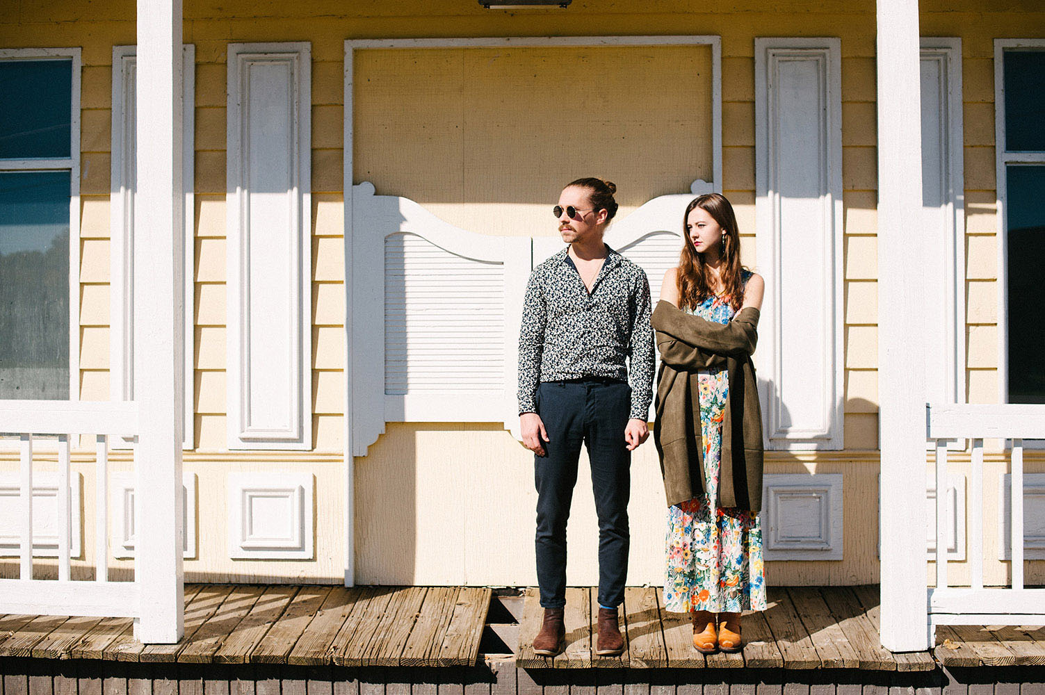Guy with man bun, round sunglasses and blue print shirt and girl with long brown hair, long floral print dress and cowboy boots in front of yellow western style building