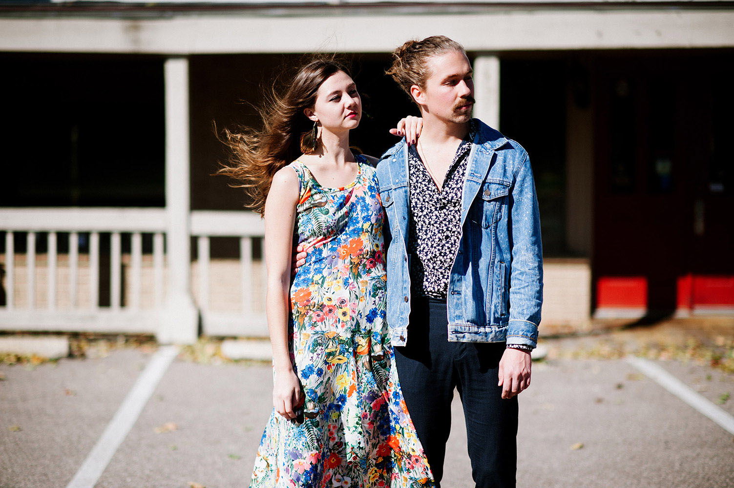 Girl with brown hair blowing in the wind, peacock feather earrings and long floral print dress and guy with man bun and jean jacket