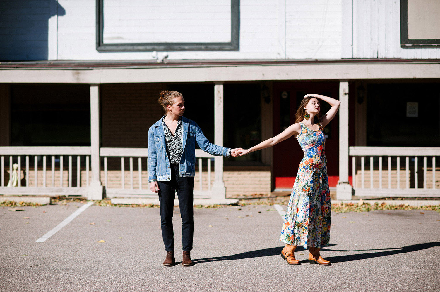 male with man bun and jean jacket and woman in long floral print dress, cowboy boots and peacock feather earrings in front of old Western building