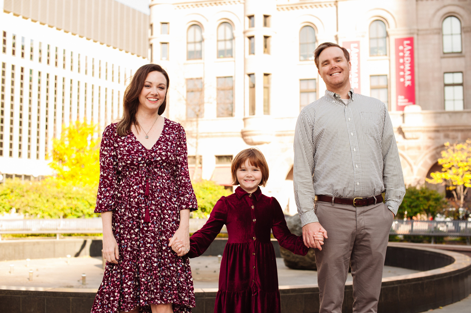 a family of 3 holding hands and smiling in front of the Landmark Center in St. Paul