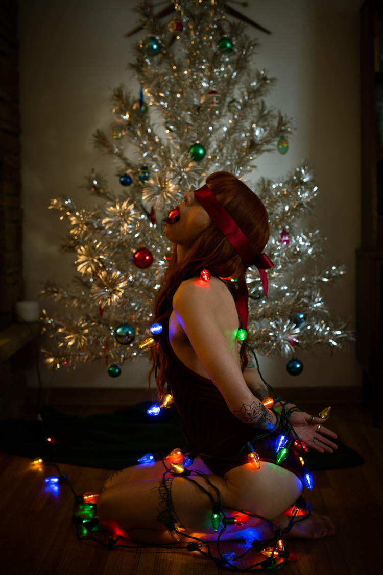 a woman in lingerie tied up in Christmas lights with a red ornament in her mouth and red tie over eyes in front of a fake silver tree